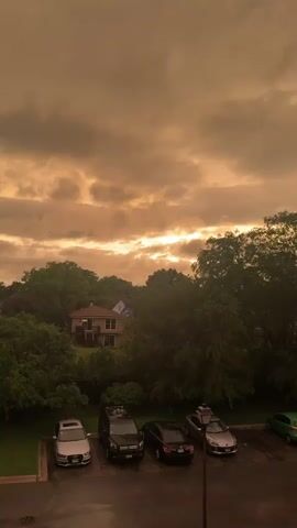 The video captures a vertical view of a residential area under a cloudy, orange-toned sky, possibly indicating sunset or a storm. In the foreground, a wet asphalt parking lot contains several parked cars: a silver SUV, a black SUV, a black sedan, and a silver sedan. Further back, nestled among lush green trees, a light-colored house with a darker roof is visible. The trees are dense, obscuring much of the background. The lighting is dim, and the clouds are heavy and textured.