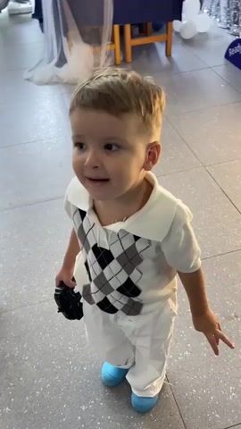 The video shows a young boy and girl in a room. The boy is wearing a white shirt with a diamond pattern, white pants, and blue shoes. He is holding a black object in his hand. The girl is wearing a pink top, a denim skirt, and pink sparkly shoes. She is holding a white stuffed animal. The room appears to be decorated with balloons, and there is a table in the background. The two children are looking at each other and appear to be interacting. The boy smiles at the camera. The girl is holding the stuffed animal and looking around. The video appears to be taken indoors.