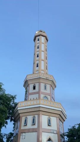 A tall, octagonal tower stands against a clear blue sky. The tower is a light cream color with decorative beige accents around the edges of each section and around the windows. Many windows are visible on the tower, some are open and some are closed, with a repeated pattern of arched windows. The tower narrows as it ascends, with a thinner, taller section at the top that also features many windows. At the very top of the tower, a small golden structure is visible. A thin wire extends from the top of the tower into the sky. Green foliage from trees is visible on the left side of the frame, partially obscuring the lower portion of the tower. The overall impression is of a unique, perhaps historic, tower against a calm sky.