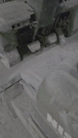 A forklift is shown covered in a thick layer of white dust. Large white fabric bags, also covered in dust, are visible in the background, some resting on wooden pallets. The environment appears to be a dusty industrial setting, possibly a warehouse or manufacturing facility.