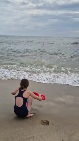 A young girl with dark hair, wearing a dark blue and patterned one-piece swimsuit, kneels on a sandy beach facing the ocean. She is holding a red toy Frisbee in her right hand. The ocean has small waves breaking on the shore, and the sky is cloudy with a muted grey and white cloud cover. Further down the beach, two adults are seated, and other people are visible in the distance. The girl is looking towards the water.