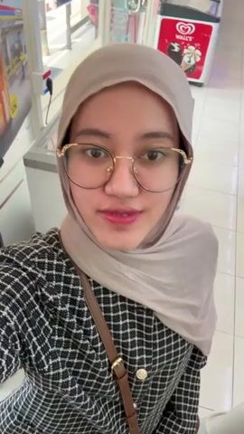 The video shows a young woman wearing a hijab and glasses. She is smiling at the camera. She is wearing a black and white checkered jacket and has a brown purse slung over her shoulder. In the background, there is a refrigerator with ice cream ads on it. The scene appears to be inside a store.