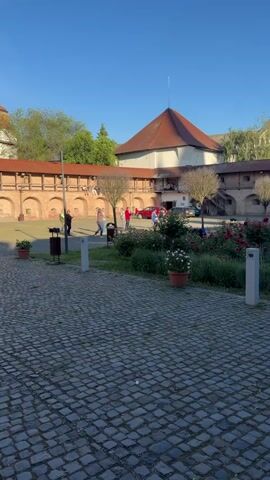 The camera shows a courtyard paved with grey stones. Several people are standing in the courtyard, near a landscaped area with bushes and flowers. The building is constructed of red bricks, with a covered walkway and arched openings on the upper level. A red car is parked in the background. Some people are on the walkway, looking down at the courtyard. The sky is clear and blue.