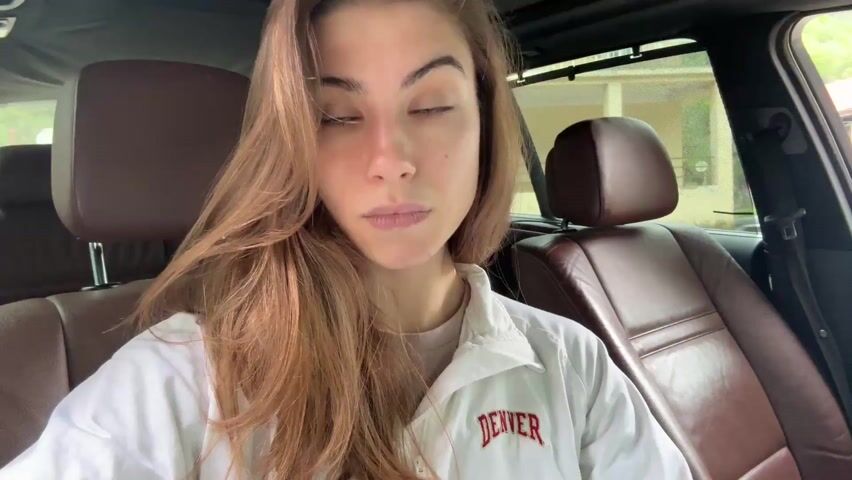 A young woman with blue-green eyes and long, light brown hair is sitting in the driver's seat of a car. She's wearing a light-colored jacket over a t-shirt. The interior of the car is brown leather. The woman is looking at the camera and talking. She holds a lit cigarette up to her mouth and smokes it.  Through the car window, we can see a building and some green foliage. The lighting is natural, and the camera is positioned close to her face.