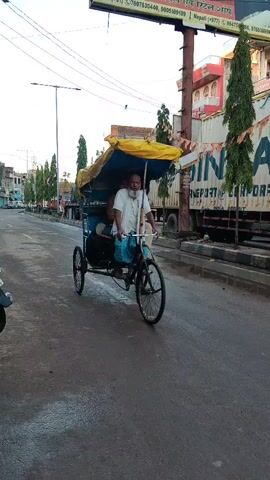 A man is driving a pedicab on a paved road. He is wearing a white shirt and blue and white checkered pants. Another man is sitting in the passenger seat of the pedicab. He is wearing a brown and orange checkered shirt and light-colored pants. There are bags in the pedicab. The pedicab has a yellow canopy. The road is wet and there are vehicles and buildings in the background.