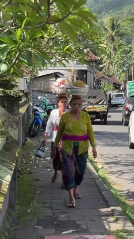 Two women walk along a paved sidewalk. Both women are carrying baskets filled with items on top of their heads. They are wearing traditional clothing, including sarongs and blouses. The woman in front is wearing a lime green blouse and a colorful sarong with a pink sash. The woman behind her is wearing a white blouse and a purple sarong with a red and white striped sash. The woman in the back is also carrying a black and white checkered bag in her left hand. They are walking on the left side of a road with traffic. In the background, there are buildings, motorcycles, cars, and lush green vegetation, including trees and mountains. The day appears to be sunny.