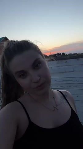 A woman is taking a selfie in front of a white wooden fence. She has light brown hair pulled back in a ponytail and is wearing a black tank top and a gold necklace. There is a building with a green roof behind her to the left. The sky behind her is a dusky orange and pink sunset. She looks into the camera and smiles.