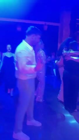 A man in a white button-down shirt and light-colored pants is dancing in a nightclub. He has blonde hair and is looking to his left with an open mouth as if singing or talking. He is raising his right hand. Behind him, several other people are dancing. The lighting is dim, with red and blue spotlights illuminating the dance floor. The background shows a dark wall with decorative lighting fixtures.