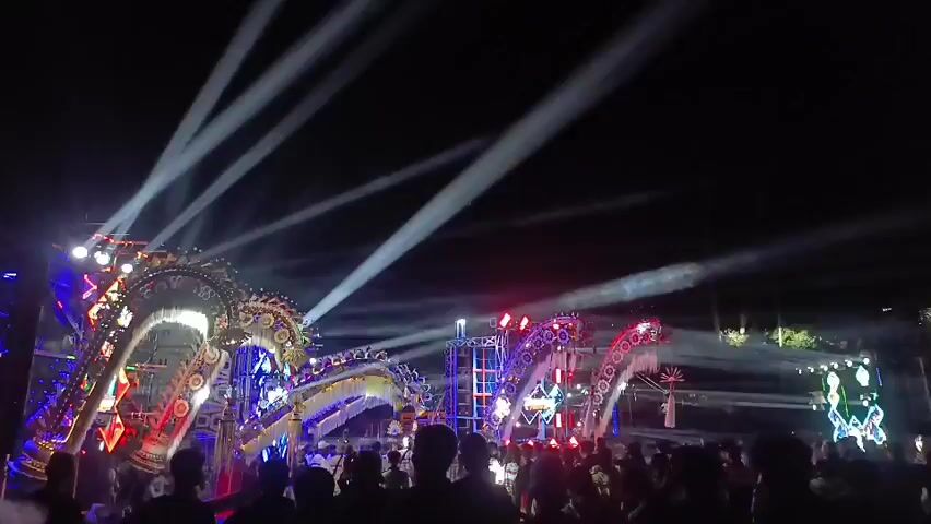 The video captures a night-time outdoor event with a large crowd gathered in front of an elaborate stage. The stage is adorned with illuminated, decorative structures resembling giant dragon or serpent heads, featuring intricate designs and vibrant LED lights in red, blue, green, and yellow. Powerful white and colored spotlights scan the dark sky, creating beams of light that cut through the darkness and illuminate the smoke or haze present in the air. The crowd, silhouetted against the bright stage, appears to be enjoying the performance. The overall atmosphere is that of a lively festival or concert.