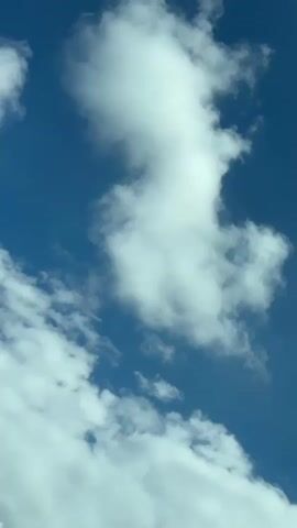 The video shows a blue sky with white clouds. A thin black wire is visible in the lower left corner of the frame. The clouds appear to be moving slowly. The sun is not visible, but the sky is bright, suggesting it is daytime.
