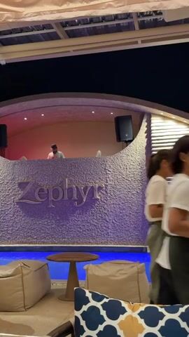 The video shows a man standing behind a DJ booth with the word "Zephyr" displayed in front of it. He appears to be a DJ and is wearing a cap. The DJ booth is situated near a pool of blue water, and there are beanbag chairs and a small table in the foreground. The environment seems to be an outdoor lounge or club at night, with artificial lighting illuminating the scene. Two other men walk by in the background, one wearing a light-colored shirt and dark pants, and the other in a white t-shirt and dark pants. A waterfall feature is visible on the right side of the DJ booth.