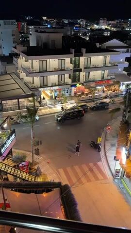 The video presents an aerial view of a bustling street in Ksamil, Albania, at night. The scene is illuminated by numerous artificial lights from storefronts, streetlights, and building facades, creating a vibrant atmosphere. Modern, multi-story buildings, primarily white with dark balconies, line both sides of the street.

Many people are visible walking on the sidewalks and in the middle of the street, which appears to be a pedestrian-friendly area or one with controlled vehicle traffic. Cars are moving slowly, with a prominent black SUV in the center of the frame and other vehicles further down the street. A scooter or motorcycle is parked near a crosswalk.

The street is lined with various businesses, including restaurants and cafes featuring outdoor seating areas, many occupied by diners. Palm trees, some illuminated with green, blue, and purple lights, are interspersed along the street. Text visible on buildings includes "L'AMO DEL MARE HOTEL" in the background and "BEST FOOD & GRILL" on a storefront. "L'Arena Beach Bar" and "L'Arena Bar Restaurant" are also visible with pink/purple lighting on the left. The overall impression is of a lively evening in a resort town.