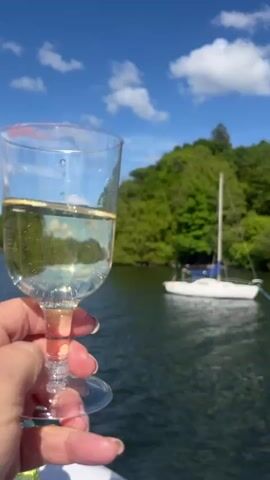 The video shows a hand holding a glass of bubbly wine against a background of a lake, green trees, and a sailboat. The sky is bright blue with puffy white clouds. The hand and glass are the foreground, with the lake and sailboat in the middle ground, and the trees and sky in the background. The wine bubbles as the narrator speaks in the audio.