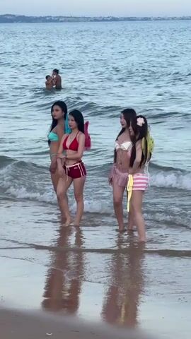 A video captures four young women in bikinis posing on a beach as waves lap at their feet. Two of the women are wearing fairy wings, one pink and one yellow. In the background, two men are visible in the water. The sky is overcast, suggesting either dawn or dusk. The sand is wet and reflects the figures of the women.