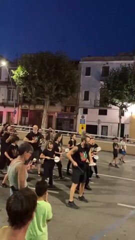 A group of people, dressed mostly in black, are performing what appears to be a street performance or parade at dusk. They are holding white circular objects, possibly tambourines or hand drums, and are moving in unison. Some individuals are wearing t-shirts with a graphic design. The background consists of buildings with lights on, trees, and streetlights. The sky is dark blue, indicating nighttime or late evening. A few spectators are visible in the foreground, including children. There are street markings on the asphalt, suggesting the performance is taking place in a street or public square. The overall atmosphere is festive.