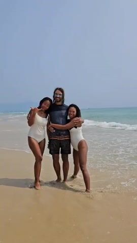 Three young adults, two women and one man, are captured standing on a sandy beach, posing for a photograph. The man, with long dark hair and a beard, stands in the center, wearing a dark grey rash guard and patterned swim shorts. He has distinctive blue face paint across his cheeks and forehead. The woman on his right embraces him, while the woman on his left stands close by.

Both women are wearing white one-piece swimsuits and are smiling broadly, making "shaka" hand gestures. The woman on the left also wears a necklace with a light-colored pendant. The woman on the right is laughing heartily. All three individuals appear happy and relaxed.

They are positioned at the water's edge, where gentle waves meet the light brown sand. The background features the expansive turquoise ocean under a clear, bright sky, indicating sunny weather. The scene depicts a cheerful moment on a beach in Bac Cam Ranh, Vietnam. There is no visible text in the image.