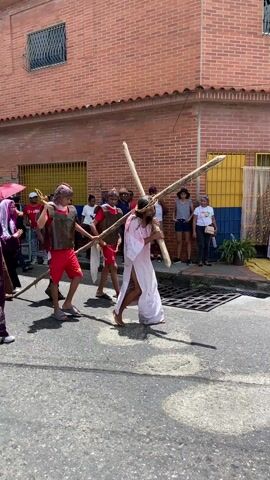 The video shows a street procession in front of brick buildings. A person dressed as Jesus, wearing a white robe with red markings and a crown of thorns, carries a large wooden cross. Several other people are dressed as Roman soldiers, wearing red clothes and helmets, and they help carry the cross. One person in a purple robe walks alongside. A crowd of onlookers lines the street, some taking photos with their phones. There's a small table with a picture on it next to a house. The day is sunny and bright.