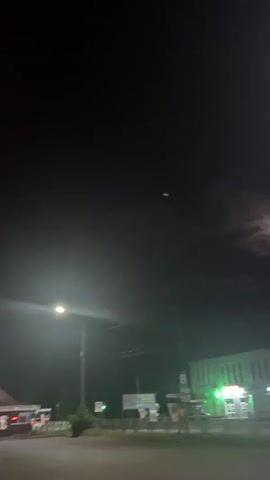A nighttime scene unfolds with a bright full moon partially obscured by clouds. The moon casts an ethereal glow, illuminating the surrounding dark sky. Below the sky, a building with green-lit windows is visible, suggesting it might be a store or gas station. To the left, a street light illuminates the foreground. The overall atmosphere is calm and serene, with the moonlight creating a sense of mystery. The time of day is clearly night. While the exact location is not specified in the visual, the context provided states Yegorlykskaya, Russia. There are no people visible in the frame, and the focus is on the celestial body and the surrounding environment.