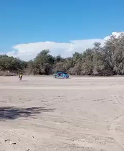 The video shows a person riding a dirt bike across a sandy, open area towards the camera. In the background, a light blue off-road buggy with multiple occupants is parked amongst sparse, bare trees. The sky is bright blue with a few scattered clouds.  The rider on the dirt bike is wearing dark clothing and a helmet. The ground shows signs of vehicle tracks. The vegetation looks dry and typical of an arid environment.
