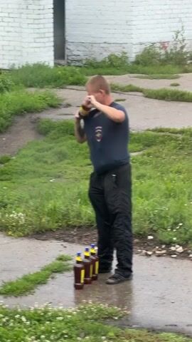A man with short blonde hair, wearing a dark blue t-shirt and black pants, stands in a grassy area with a paved path. He is holding a brown bottle with a yellow cap and appears to be drinking from it. In front of him, on the paved path, are four identical brown bottles with yellow caps standing upright in a line. The man is positioned facing the bottles, and the scene appears to be outdoors, with a white brick building in the background.