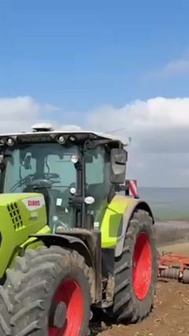 The video shows a green and red CLAAS Arion 660 tractor working in a field on a sunny day with a blue sky and scattered clouds. The tractor is pulling a piece of farm equipment, likely a disc harrow or roller, across the brown, tilled soil. The landscape in the background includes rolling hills, trees, and a mix of agricultural and natural areas. The focus is primarily on the tractor and its operation in the field.