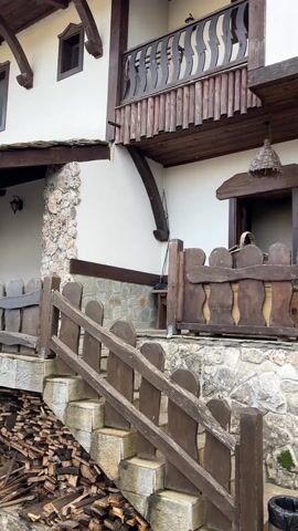 The camera slowly pans along the facade of a building with rustic, old-world charm, starting from a ground-level view of stone steps and ascending towards the building's porch. The building itself is a mix of whitewashed walls and rough-hewn stone, with exposed wooden beams adding to the rustic aesthetic. The porch is surrounded by a wooden fence with a distinctive scalloped design. Strings of what appear to be dried red peppers are suspended from the eaves, adding a splash of color. An old, repurposed wooden tool stands as a decorative element near the fence. Transitioning to the right, the camera reveals a wooden pergola with a small, old-fashioned lantern attached to one of its support posts. Underneath the pergola, there's a glimpse of outdoor seating, suggesting a patio area. The video is devoid of people, and the focus remains on the architectural and decorative details of the building and its surroundings.