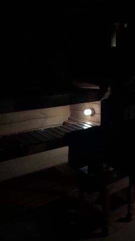 The video depicts the interior of a dimly lit room, possibly a sauna or wooden structure. A small, bright light illuminates a portion of the wooden bench and wall. Another wooden structure resembling a small table or step stool is partially visible in the foreground. The rest of the room is shrouded in darkness, and the overall atmosphere is dark and indistinct. The video is static with no movement. Sounds of Russian speech are present.