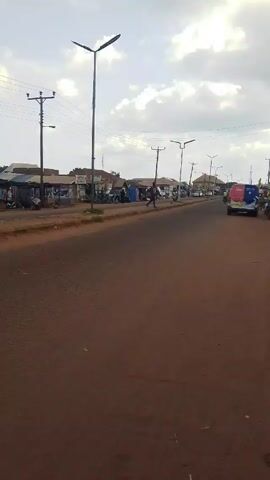 A handheld camera captures a scene from a slightly elevated position, looking down a road in what appears to be a small town. The road surface is a reddish-brown color. On the left side of the road are small shops or stalls. People can be seen walking around and interacting with the stalls. Power lines stretch across the sky above the road. A man walks across the road, and a colorful van is parked on the right side. In the background, more buildings and trees can be seen. The sky is cloudy, suggesting overcast weather.