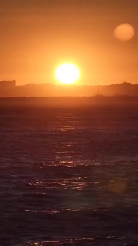The video shows a wide shot of a body of water at sunset. The sun is a bright, large orb in the sky, casting a warm orange glow over the scene. The water is choppy with small waves. In the distance, a silhouetted cityscape can be seen against the sunset. There are no people or specific objects visible, but the overall impression is one of natural beauty and tranquility.