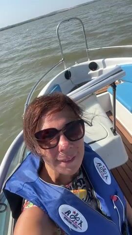 A woman wearing sunglasses and a blue life vest smiles at the camera from a moving boat. Her hair is blowing in the wind. The boat is on a body of water with a distant shoreline under a clear blue sky.