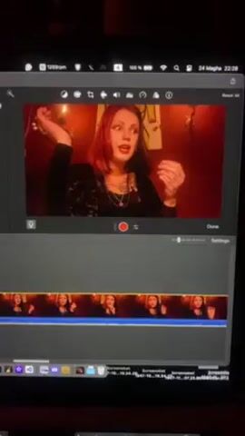 A woman with red hair is displayed on a MacBook Pro screen. She is wearing a black top and several necklaces, and is using her hands to express herself. The video is in a recording and editing stage, as it is shown in the iMovie application. The background is a dim red color, possibly from lighting. There is a thumbnail strip of the video, at the bottom of the screen. The computer's system bar shows the date, time, and battery level.