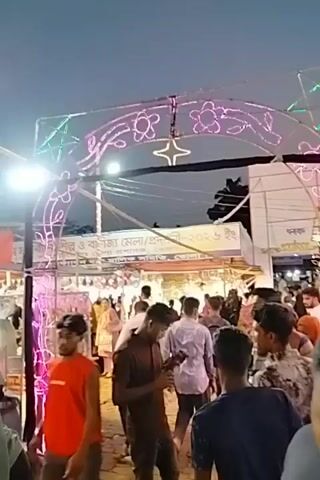 This video captures an evening scene at an outdoor market or fair, likely in Charfasson, Bangladesh, as suggested by the context. Numerous people, predominantly adults, are seen walking through the bustling area. Overhead, an elaborate display of colorful LED lights forms arches and rectangular patterns, illuminating the space. A tall palm tree, wrapped in white string lights, stands out against the dark blue sky.

The market features various stalls or booths, mostly covered in white tarps or structures. A body of water, possibly a pond or canal, is visible in the foreground, reflecting some of the lights and structures. People are walking along a path or bridge adjacent to this water feature.

At the beginning of the video (0:00-0:01), a banner with Bengali text is partially visible, appearing to include "মেলা" (fair) and "বাণিজ্য" (commerce), suggesting it might be a trade fair or general market. Another sign with Bengali text, possibly "মেহরাব" (Mehrab), is briefly seen on a booth in the background around 0:04-0:05. The overall atmosphere appears lively and festive, characteristic of an evening event.
