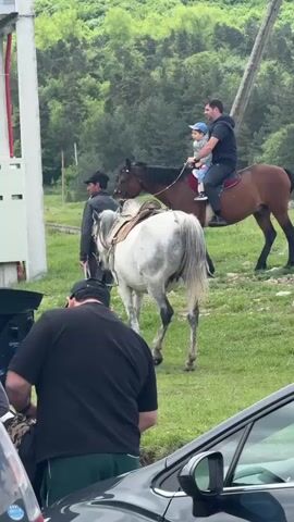 The video shows a man riding a brown horse with a child sitting in front of him. Another man is holding a white horse. The riders are on a grassy area with trees in the background. A car is parked nearby, and people are standing around the car. The scene appears to be taking place in a rural area.