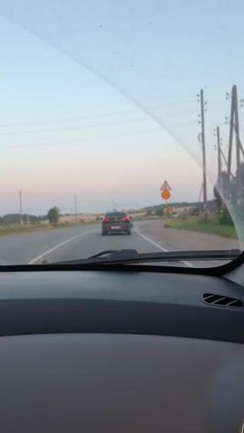 The video shows the view from inside a car driving on a two-lane road. The car's dashboard is visible in the foreground, with a grey textured panel and a circular air vent. The windshield is slightly dirty. Ahead, another car is driving in the same direction. There are trees and fields on either side of the road, and power lines are visible in the distance. Traffic signs are also present, including a triangular warning sign and a circular speed limit sign indicating 40. The sky is a pale blue with hints of pink, suggesting it might be late afternoon or early evening. The camera is pointed forward, capturing the road ahead and the car in front. The video is taken from a passenger's perspective or from the driver's seat.