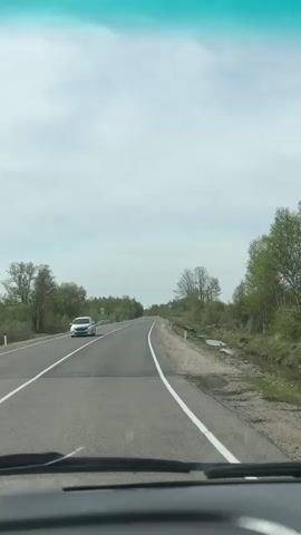 The video shows a car driving on a road with two lanes and white lines marking the lanes. Trees line both sides of the road. Another car is seen driving in the opposite direction. The sky is overcast. The video then shows a pair of feet in white socks resting on the dashboard of the car.