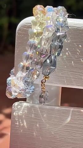 The video shows a close-up of a beaded bracelet made of clear and blue faceted beads. The bracelet has a gradient effect, transitioning from clear to a light yellow, then to a light blue, and finally to a darker blue. A small, gold-colored charm hangs from a chain attached to the bracelet, featuring a clear, faceted bead shaped like a small gem. The bracelet is resting on a white surface, possibly a piece of furniture or a decorative item, which appears to be outdoors given the sunlight casting shadows and reflections. The background is blurred, suggesting a shallow depth of field, with hints of green foliage visible, indicating an outdoor setting. The lighting is bright, highlighting the sparkle and facets of the beads.
