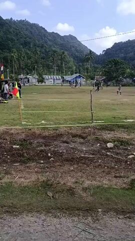 The video shows a soccer game being played on a field with a natural grass surface. The field is marked with white lines. In the background, there are trees and hills. There are several players on the field, wearing dark colored uniforms. One player is seen running with the ball, while others are positioned around the field. There is a flag on a pole, possibly marking a corner of the field. A person, possibly a spectator, is visible in the foreground, looking towards the game. The sky is partly cloudy. The overall impression is that of a casual soccer match in a rural or semi-rural setting.