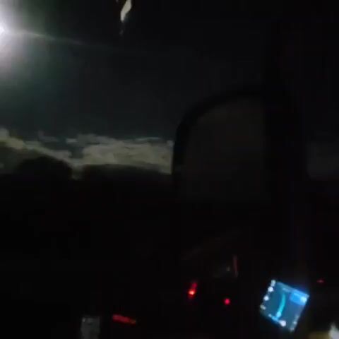 The video displays a nighttime scene from inside a vehicle, looking through the windshield. A bright, square light source, possibly a street light or a high beam, illuminates the dark sky, creating a lens flare effect. Below this light, a layer of clouds is visible, appearing lit from above. The foreground is dark, with some faint red and blue lights suggesting dashboard instruments or electronic devices within the car. A screen, possibly a GPS or phone, is also visible in the lower right corner, displaying a map interface. The overall impression is of a night drive with limited visibility due to darkness and the bright light in the sky.