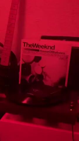 A record player is playing The Weeknd's "House of Balloons" album. The image is lit with red light. A black guitar leans against the wall in the background, and a speaker is visible behind the record player.