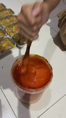 The video shows a close-up shot of a person's hand holding a wooden utensil and stirring a thick, orange sauce in a clear plastic container. The container is filled with the sauce, and the utensil is creating swirls and ripples in it. In the background, several foil containers filled with dim sum are arranged in rows on a tiled floor. The lighting suggests an indoor setting, likely a kitchen or food preparation area.