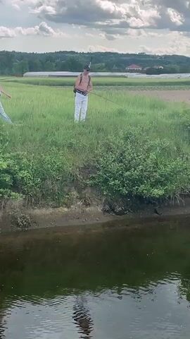A video shows three people standing on a grassy bank overlooking a narrow body of water. One person is a young woman in a pink top and jeans. The other two people appear to be young men, both shirtless and wearing hats. One of the young men is wearing white pants and carrying a fishing rod slung over his shoulder. The other young man is wearing jeans and carrying a backpack. The people are standing on a green grassy bank with bushes. On the other side of the bank is a green field. In the background, there are more fields and some buildings. The sky is cloudy.
