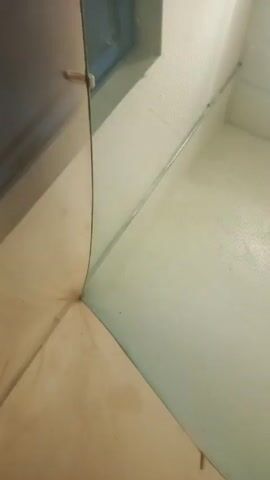 The video shows a close-up, angled shot of a partially obscured, potentially indoor corner, showing a floor, a wall, and a screen or filter. The floor appears to be beige tile or linoleum and has some marks or discoloration. The wall is a light color and appears to be painted or wallpapered. A metallic or reflective trim piece runs along the seam between the wall and the screen. The screen or filter is translucent with a visible mesh pattern. The corner forms a sharp, defined edge. Obscuring a portion of the left side is a dark, blurred surface that is likely a cabinet or other piece of furniture.