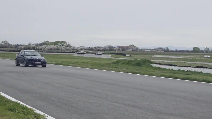 The video shows a dark blue BMW racing on a paved racetrack. The car passes by the camera, and then the camera follows it as it drives away. There is a sign in the background that says "MAFRA cosmetics for your car". In the final shot, a man's head is visible in the foreground as he watches the car drive away. The sky is overcast, and there are trees and mountains in the distance.