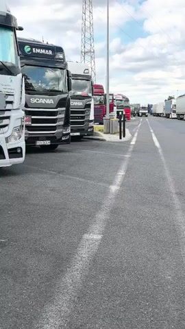 This media file shows a line of parked semi-trailer trucks in a large asphalt lot, likely a rest stop or truck yard. The trucks are predominantly white and silver, with several brands visible, including Scania and EMMAR. The scene appears to be outdoors during the daytime, with a cloudy sky and visible power lines and tall transmission towers in the background, suggesting an industrial or highway-adjacent location. The perspective is from ground level, moving forward along the paved area between lanes of parked trucks. There is no visible activity of people in the frame.

The trucks are mostly facing away from the camera, with their rear trailers visible. Some trailers have markings, including one with the text "NOTHING ELSE BUILDS PERFOR." The lighting suggests it's a bright, overcast day. The presence of multiple large trucks parked in an organized manner indicates a location where commercial vehicles congregate, such as a truck stop, a logistics hub, or a border crossing. The overall impression is one of a functional, commercial transportation environment.