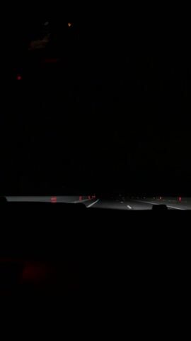 The video, shot from inside a moving car at night, shows a dark, four-lane highway receding into the distance. The road surface is visible, marked with white lane dividers and flanked by guardrails. Small red lights are attached to the guardrails, presumably for nighttime visibility. The car's dashboard is visible in the foreground, obscuring the view below the windshield. The overall scene is dimly lit by the car's headlights and distant road lights. There are no other cars visible in the immediate vicinity.