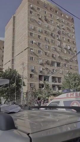The video shows a tall, multi-story apartment building with visible damage. Several windows are broken or missing, and there are large openings in the structure, suggesting a collapse or explosion. An ambulance with "EMERGENCY" written on its side is parked in front of the building, and several people in uniforms, possibly emergency responders, are gathered near the ambulance and the damaged area. Trees and power lines are also visible in the scene. The view is partially obstructed by the roof of a vehicle in the foreground.