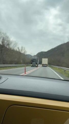 A car drives on a road between mountains on an overcast day. The roadway has red stripes and red traffic delineators. Two trucks, a car, and a utility vehicle are in front of the car that the video is taken from. It appears to be raining.