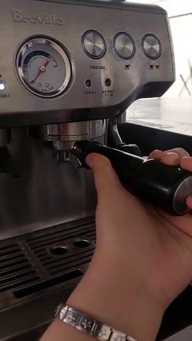 A person with a silver charm bracelet secures a black portafilter into a Breville espresso machine. The machine has several buttons, a pressure gauge, and a stainless steel drip tray. The environment appears to be indoors, with a glimpse of an outdoor area visible in the background.
