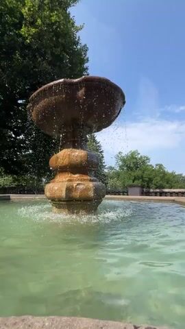 A fountain sprays water into a pool, surrounded by trees and a blue sky with some clouds. The fountain is made of stone and has a classic design. The water is clear and reflects the sky. There are no people visible in the video. The environment appears to be a park or garden.