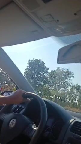The video begins inside a car, showing the dashboard and a rearview mirror with a cross hanging from it. The car appears to be moving along a road with trees on either side and a clear blue sky. In the foreground, a driver's hand is visible on the steering wheel. The view changes to show the exterior of the car, with a police officer in uniform and a high-visibility vest standing on the side of the road and waving. Two other individuals are also present on the roadside. The camera then shifts to a side view of the car as it continues to move along the road, showing more trees and vegetation.